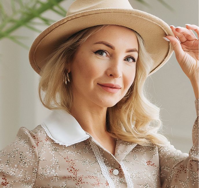 Woman in hat smiling indoors, wearing floral blouse.