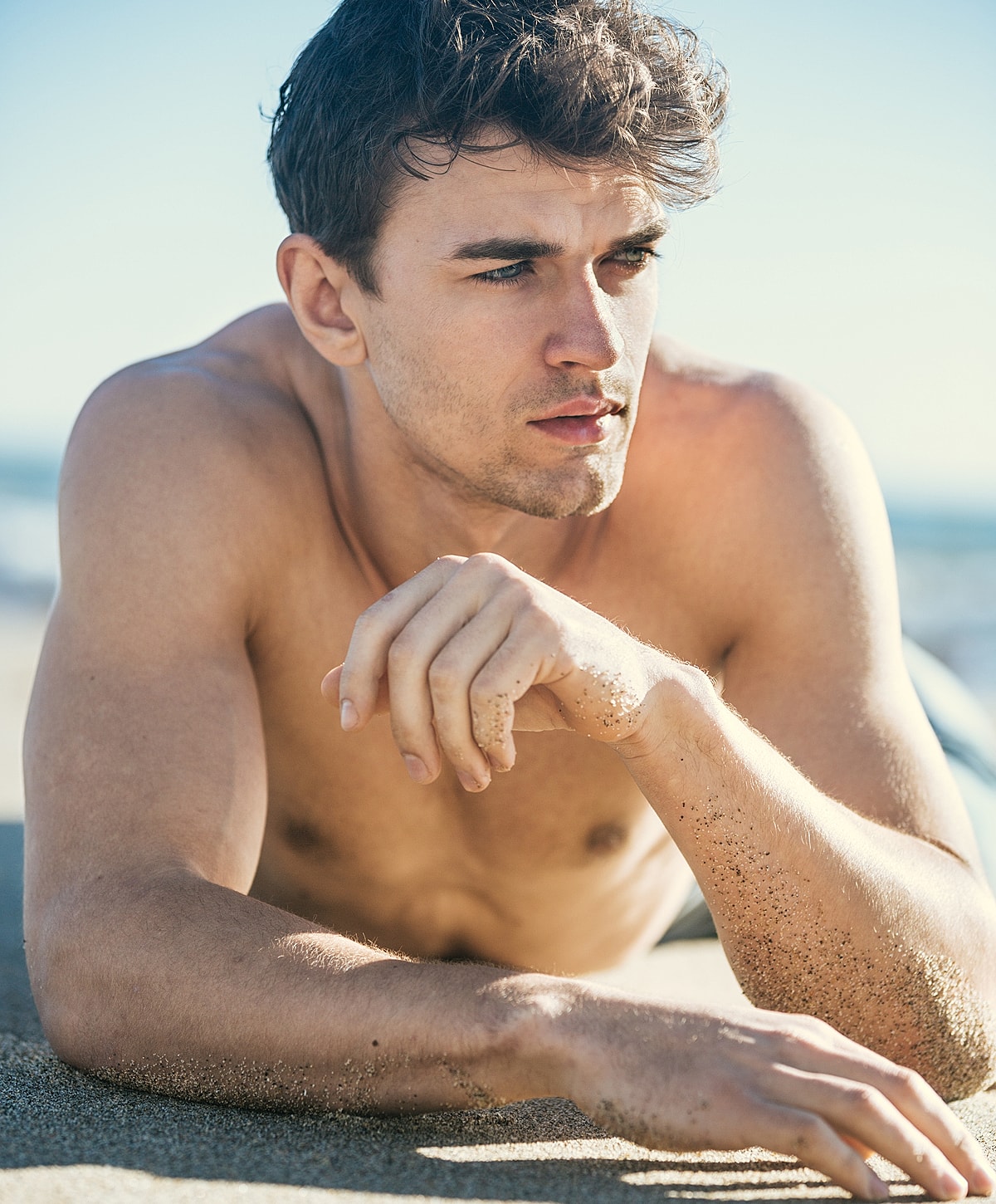 Shirtless man relaxing on sandy beach.